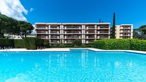 Ein Swimmingpool mit glasklarer Wasseroberfläche, umgeben von einer gepflegten Hecke, und ein mehrstöckiges Gebäude mit Balkonen im Hintergrund.