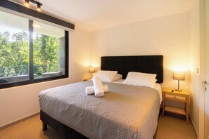 A neatly made bed with a headboard, two bedside lamps, and a window with a view of trees.