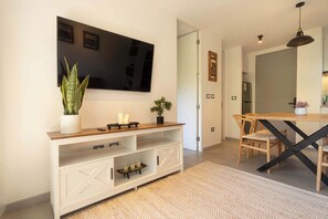 A modern living room with a flat-screen TV, a wooden console table, a dining area with a wooden table and chairs, and a patterned rug.