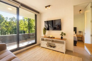 A modern living room with a large sliding glass door leading to a balcony, a white entertainment unit with a flat-screen TV, and a cozy seating area.