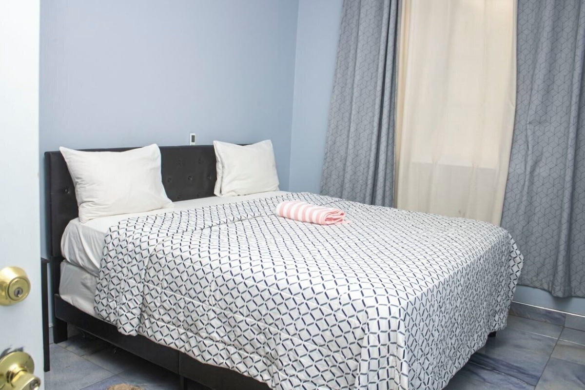 Una cama individual con una colcha estampada y dos almohadas, una puerta con picaporte de latón y una ventana con cortinas.