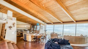 Un'ampia zona giorno con soffitto in legno, una grande finestra vista oceano, un'area pranzo e una cucina con elettrodomestici moderni.