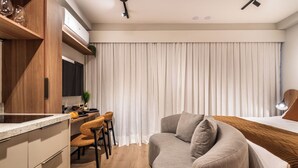 A modern hotel room with a kitchenette, a sofa, a dining area, and a large window with curtains. - StayHome Allianz Park Queen Bed Varanda (São Paulo)