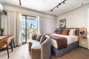 Egy modern szállodai szoba, amelyben egy nagy ágy, egy íróasztal székkkel, egy kanapé és egy városra néző kilátással rendelkező erkély található.