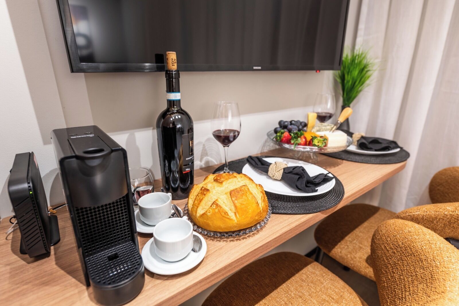 A hotel room with a wooden table set for breakfast, including bread, wine, and fruit.