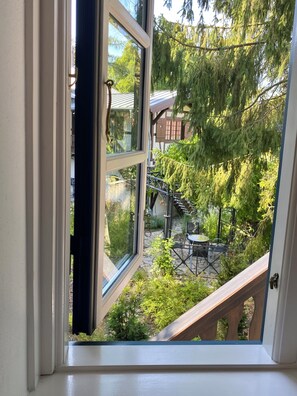 Une fenêtre donnant sur un jardin, avec notamment une terrasse en bois et du mobilier d’extérieur.