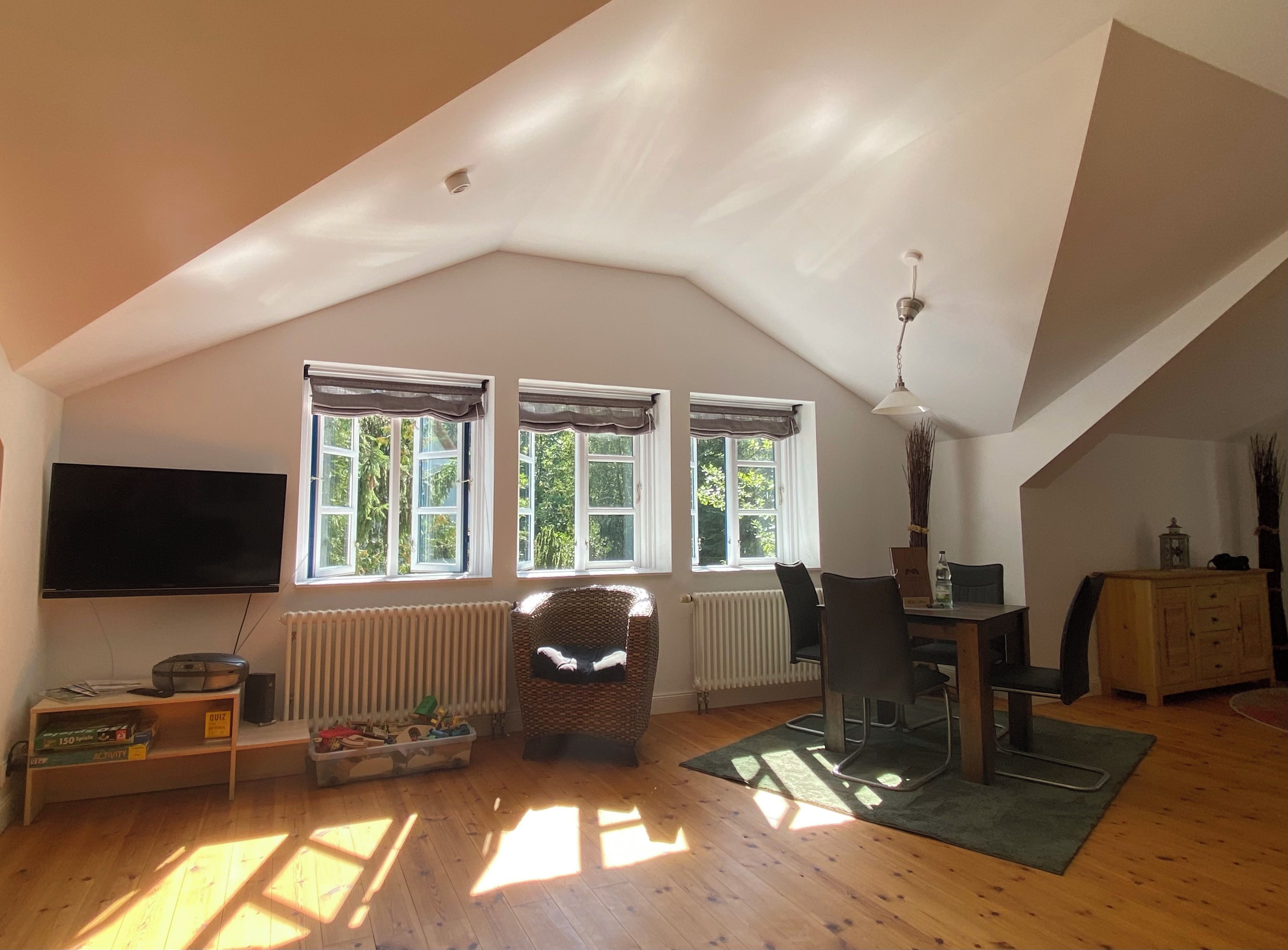 A spacious attic room with a sloped ceiling, a flat-screen TV, a dining table with chairs, and a radiator.