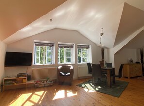 Ein geräumiges Dachzimmer mit geneigter Decke, einem Flachbildfernseher, einem Esstisch mit Stühlen und einer Heizung.