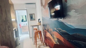 A room with a mural, a television mounted on the wall, a wooden table, two stools, and a bed with a patterned bedspread. - Lovely studio in mas provençal with private parking (Avignon)