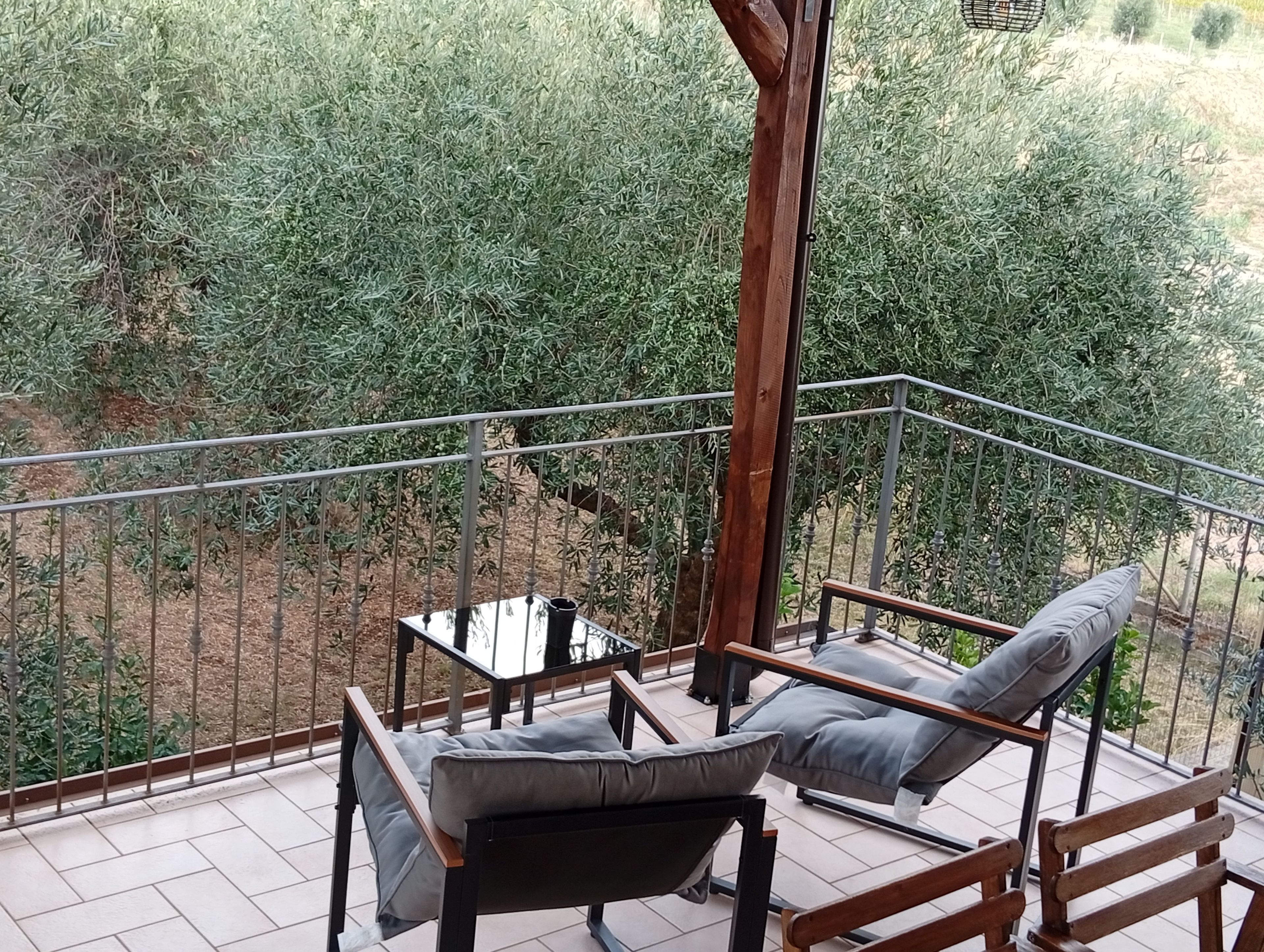 A balcony with a view of dense greenery, a wooden chair, a sofa with cushions, and a metal railing.