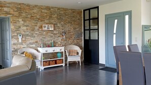 Un salon avec un mur en pierre, une console blanche, un fauteuil en osier, un canapé et un coin repas avec des chaises et une table.