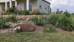 Une maison à deux étages dotée d’un patio, entourée d’un jardin aménagé avec différentes plantes et fleurs.