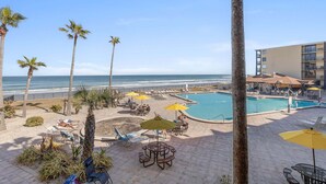 Área de piscina com espreguiçadeiras e guarda-sóis, vista para a praia e um edifício com varandas.
