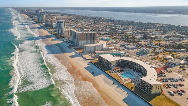 An aerial view of a coastal city with a beachfront hotel featuring a pool, parking, and a view of the ocean. - Hawaiian Inn 502 (Daytona Beach Shores)