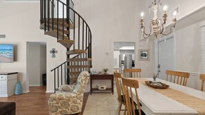 A dining area with a long table, wooden chairs, a chandelier, a spiral staircase, and a patterned armchair. - Sea Woods Villa 277 (New Smyrna Beach)