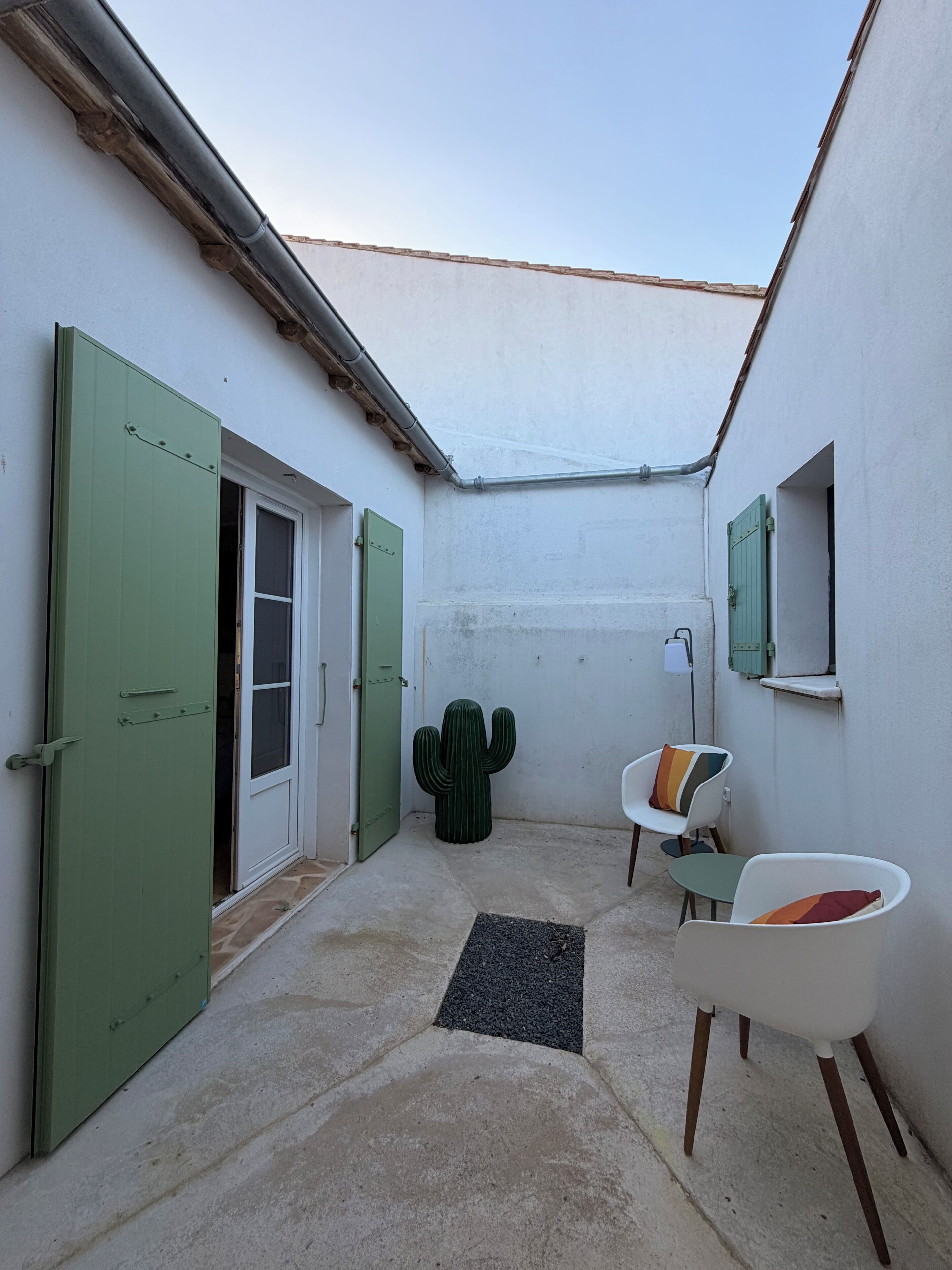 A narrow courtyard with a cactus, a white chair with a colorful cushion, and a green door.