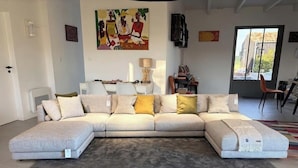 A modern living room with a sectional sofa, a coffee table, and a large painting on the wall. - L'ARS DE VIVRE – Exceptional Home in Ars-en-Ré | Pool | Sleeps 10 (Ars-en-Ré)