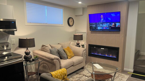 Una sala de estar moderna con un sofá, una mesa de centro de vidrio, una chimenea, un televisor montado en una pared de madera y una zona de cocina con estufa.