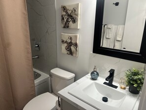 Une salle de bain comprenant des toilettes, un lavabo et une douche. Deux œuvres d’art sont accrochées au mur, et un miroir se trouve au-dessus du lavabo.
