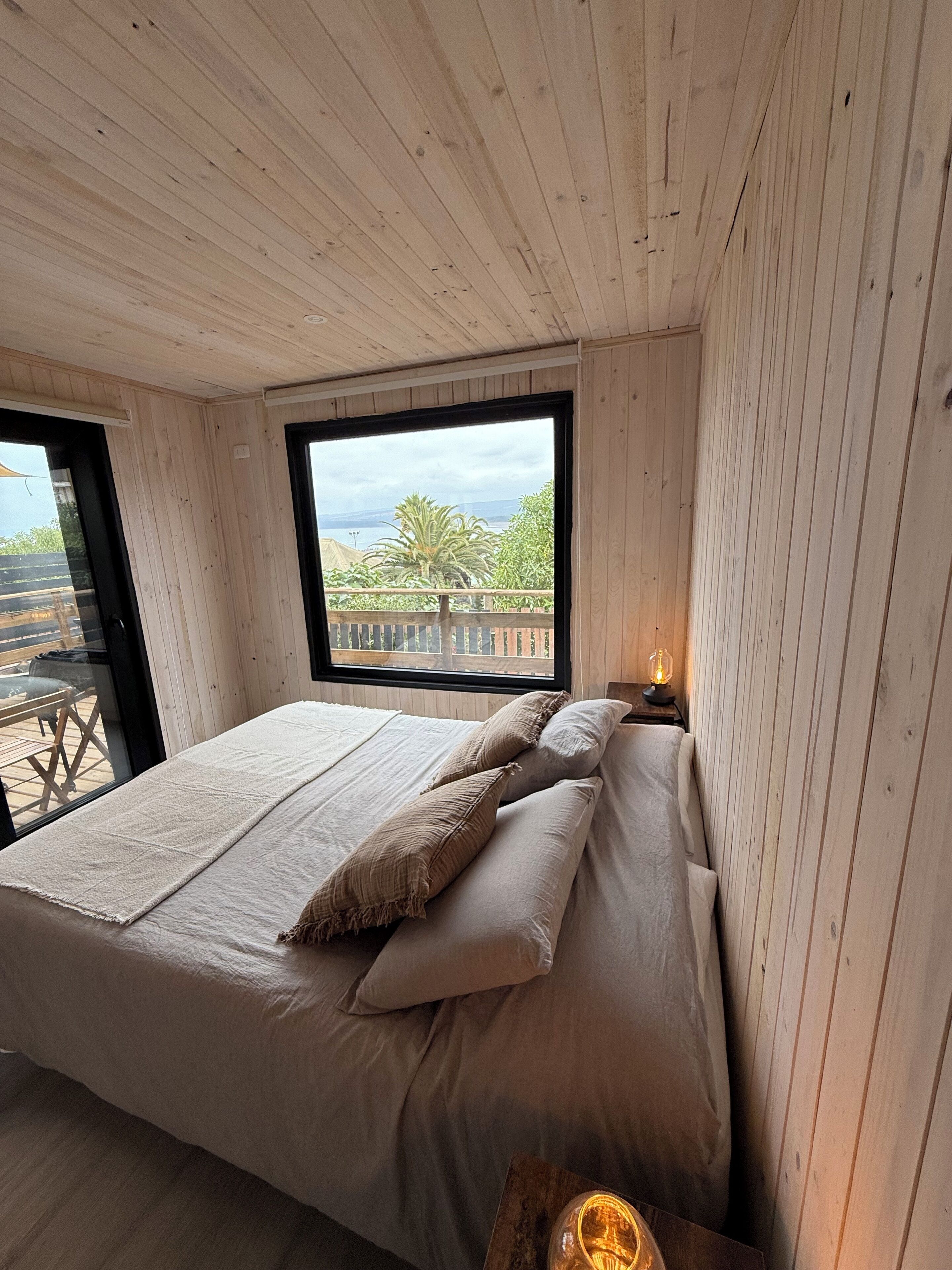 A bedroom with a large bed, wooden walls, a window with a view of greenery and a distant landscape.