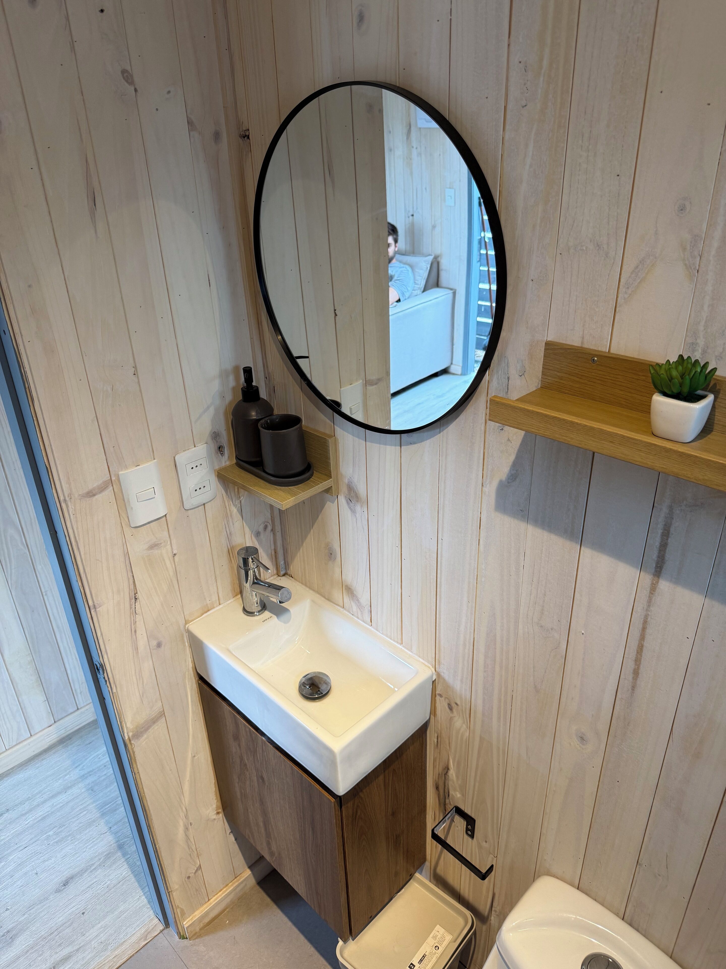 Un baño con pared de paneles de madera, un espejo redondo, un lavamanos blanco, un inodoro y una estantería con una planta.