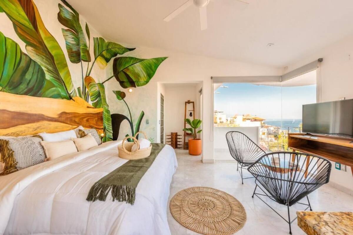 Un dormitorio con cama, televisión, balcón con vistas y una planta grande.
