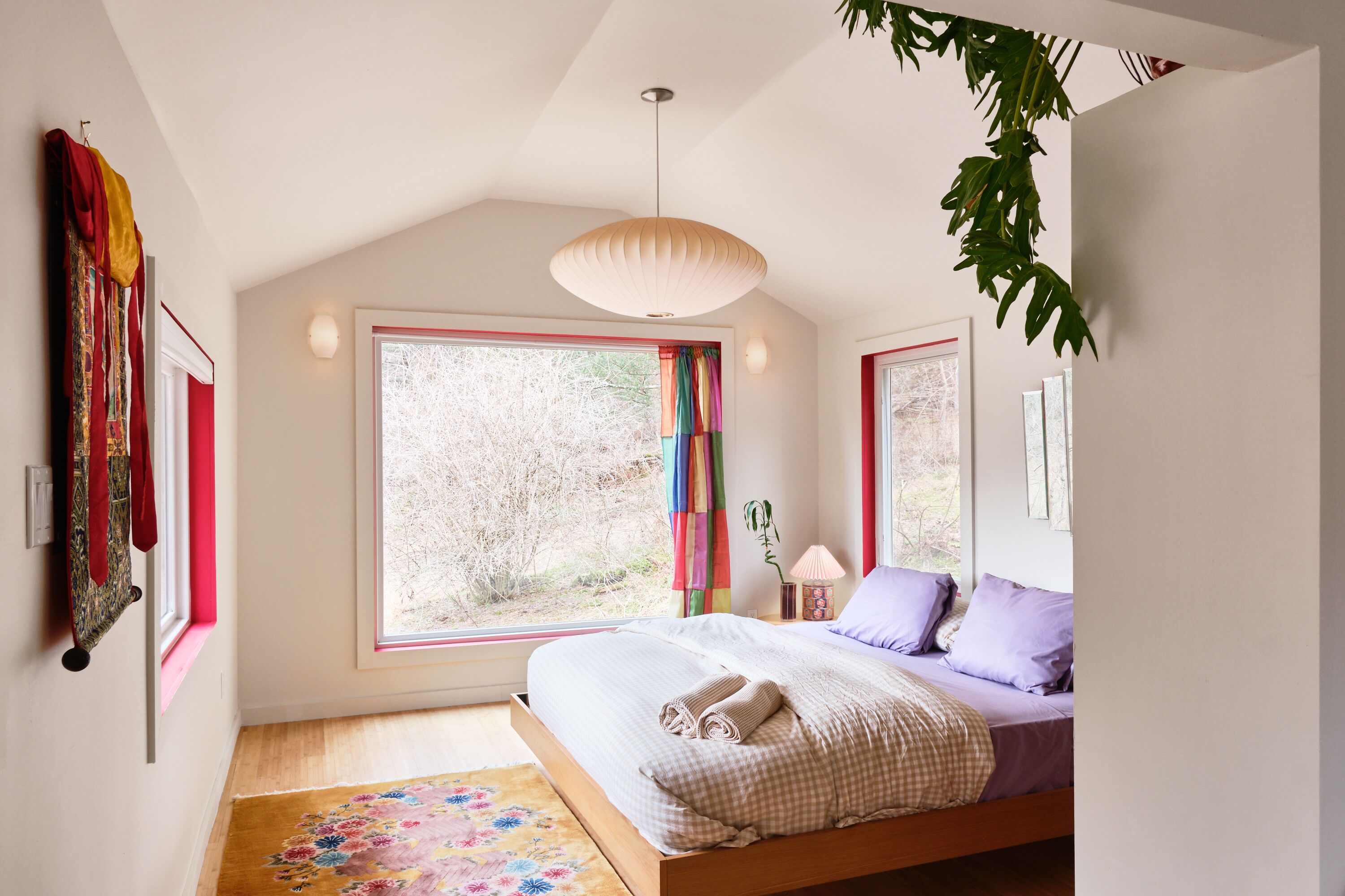 A bedroom with a large window, a bed, a hanging plant, and a light fixture.