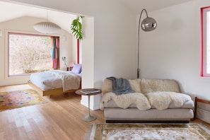 A bedroom with a bed, a sofa, a small table, a floor lamp, and a large window.