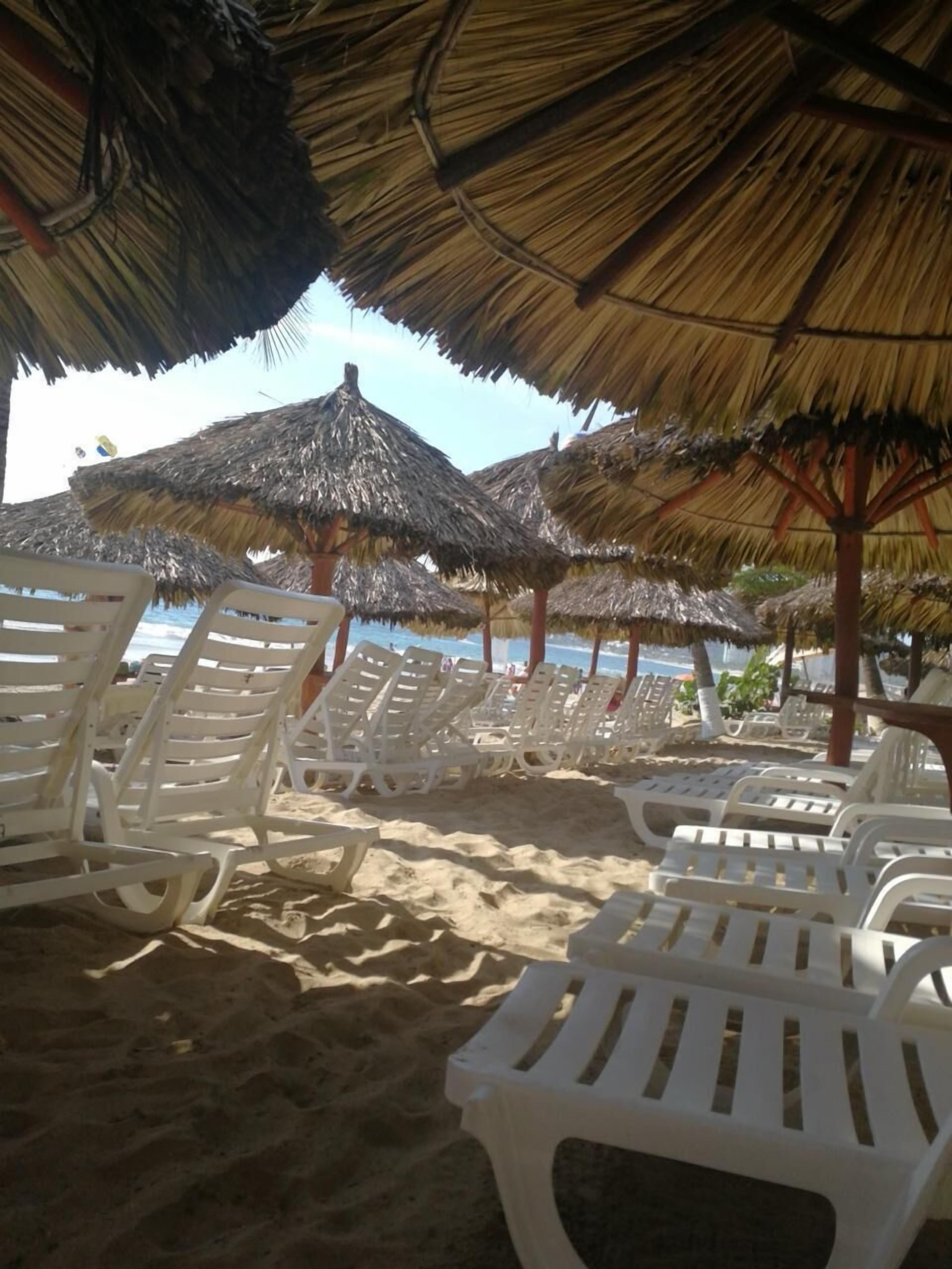 Uma praia com guarda-sóis de palha e cadeiras de plástico brancas.