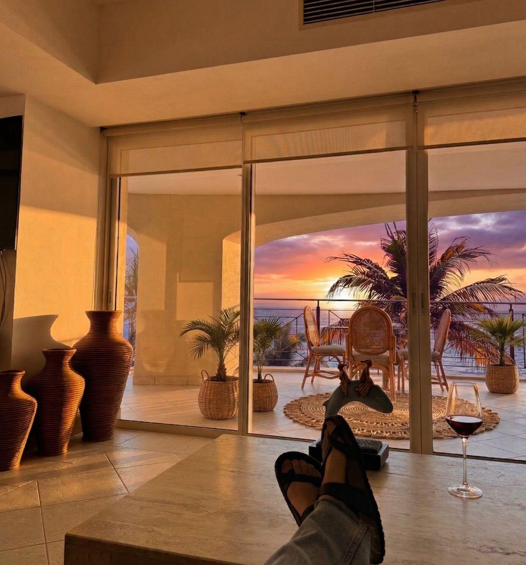 Ein Zimmer mit einer großen Glastür, die auf einen Balkon mit Sonnenuntergangsblick führt, mit Korbmöbeln und einem Weinglas.
