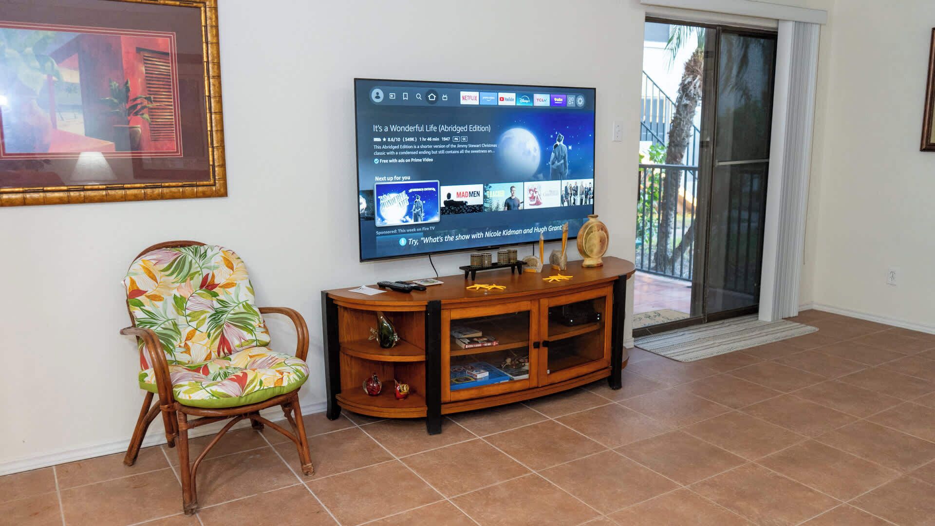 Un salon avec un téléviseur diffusant un film, un meuble de rangement en bois, un fauteuil à motifs floraux et une porte-fenêtre coulissante donnant sur un espace extérieur.