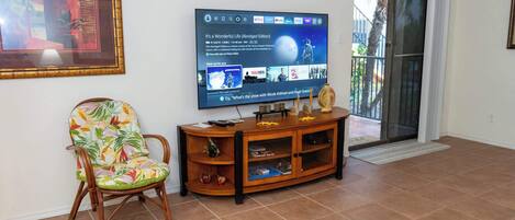 A living room with a television displaying a movie, a wooden entertainment center, a floral chair, and a sliding glass door leading to an outdoor area.
