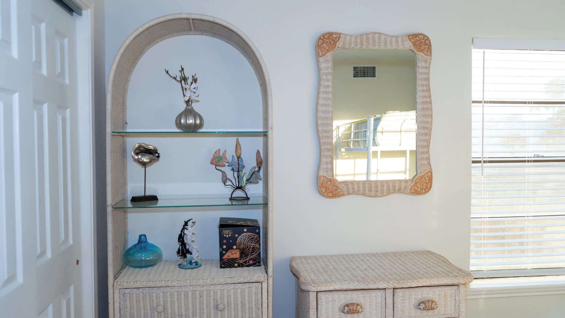 Une pièce comprenant une étagère en verre où sont exposés des objets décoratifs, un miroir avec un cadre en osier et une commode en rotin.