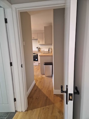 A hallway leading to a kitchen with wooden flooring, white cabinets, and a small trash bin. - Cozy 1 bed Annex (Coleorton)