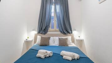 Ein Schlafzimmer mit Bett, Vorhängen, Kissen, einem Nachttisch mit Lampe und gerollten Handtüchern.