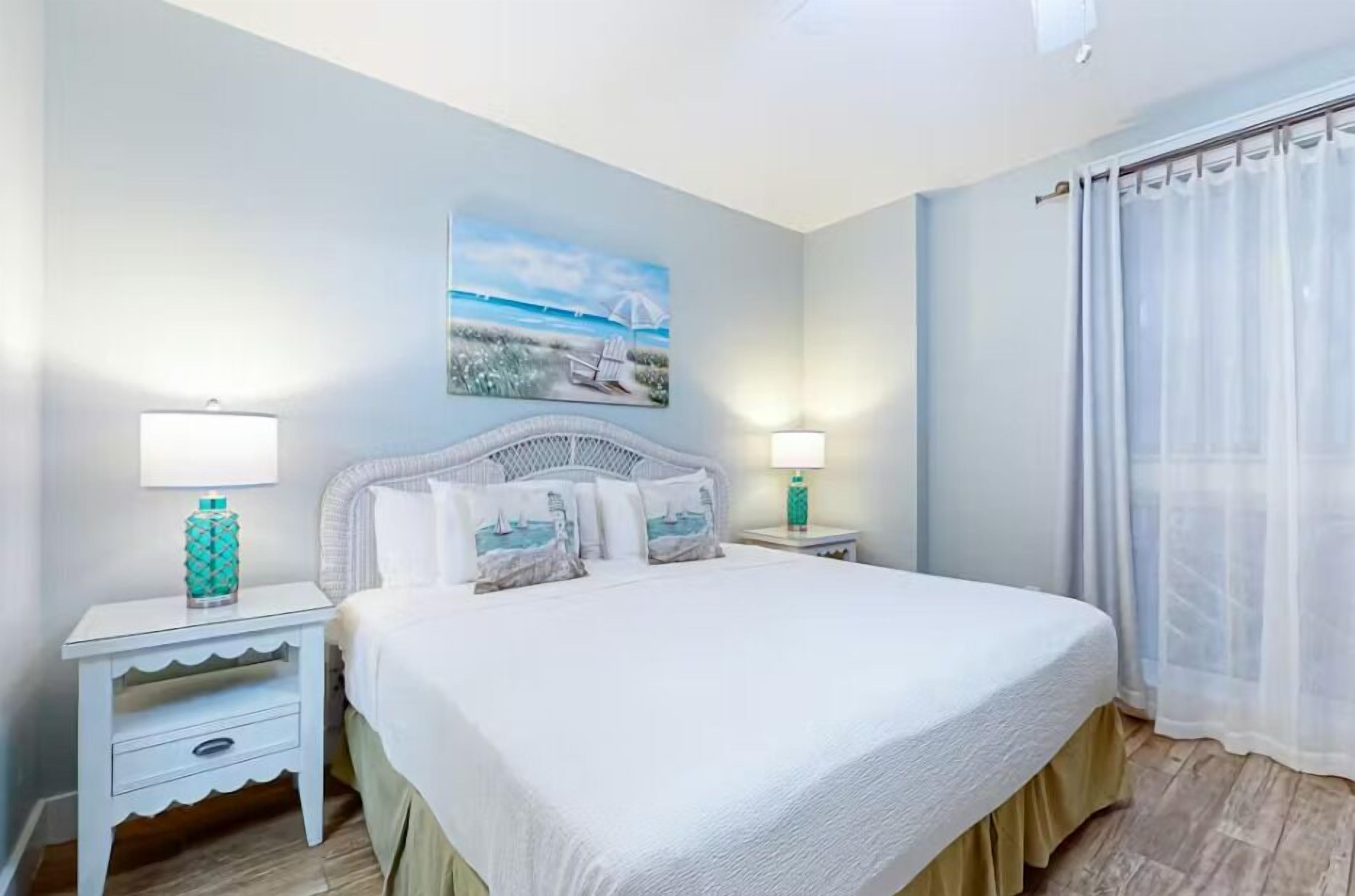 Een slaapkamer met een groot bed, twee nachtkastjes met lampen, een schilderij aan de muur en een raam met een doorschijnend gordijn.