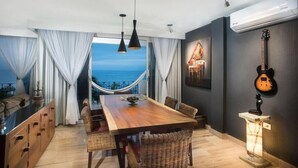 Un espace repas avec une table en bois, des chaises en osier, des luminaires suspendus et une vue sur l’océan.