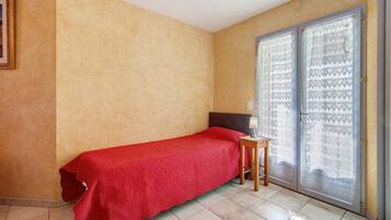 Een enkel bed met een rode sprei in een kamer met beige wanden en een betegelde vloer.
