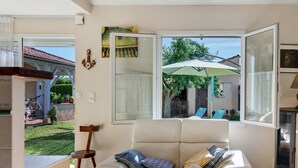 A living room with a sofa, a dining table, and a view of the garden through a large window. - Gîte la Cache-maille in Bias (Bias)