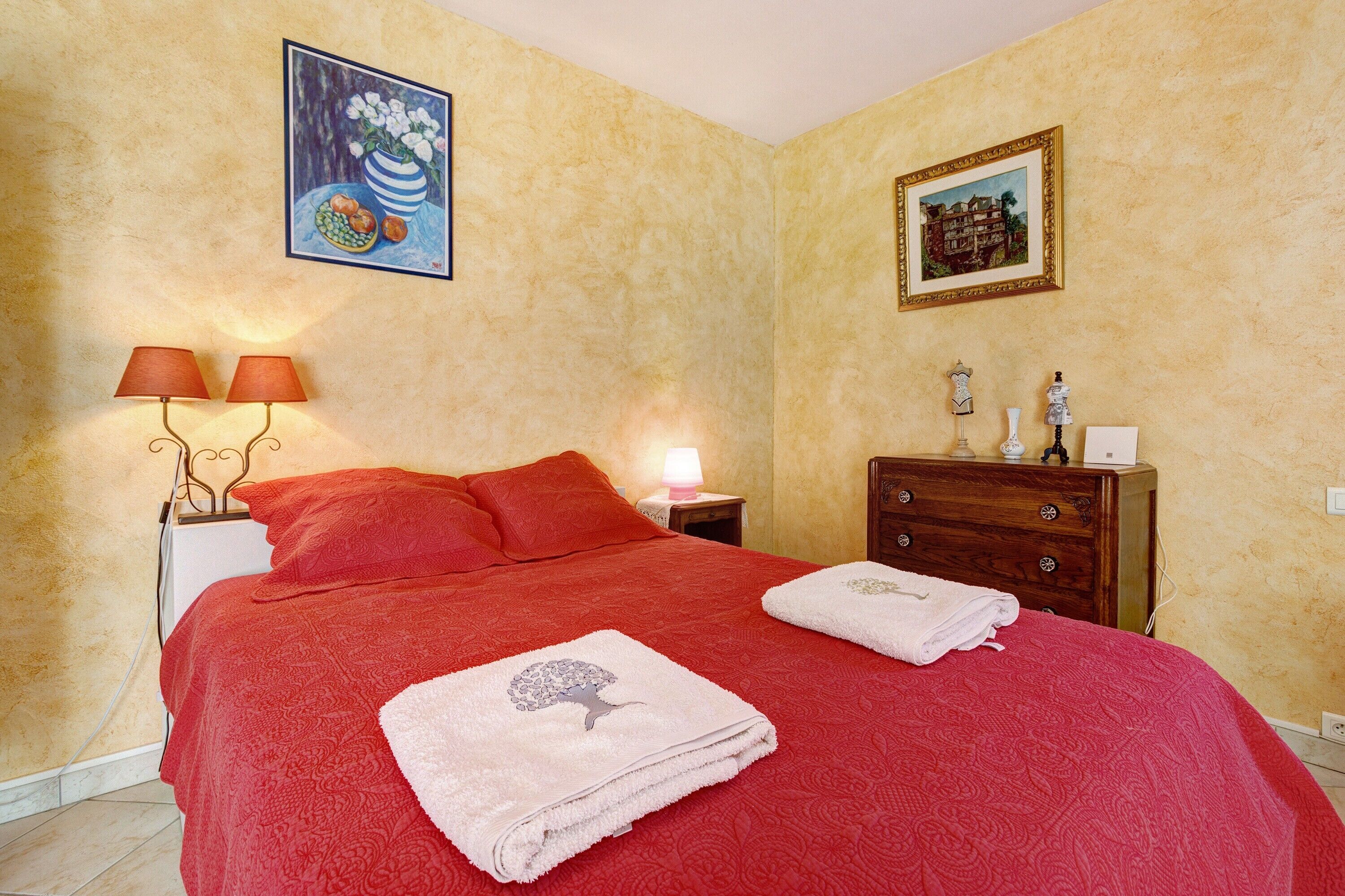 A bedroom with a red bedspread, two pillows, a nightstand with a lamp, a dresser, and two framed pictures on the wall.