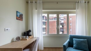 A room with a desk, chair, and a blue sofa. There is a window with curtains, a painting on the wall, and a view of buildings outside. - Maya Casa Verde - Premium Studio in Central Rome (Rome)