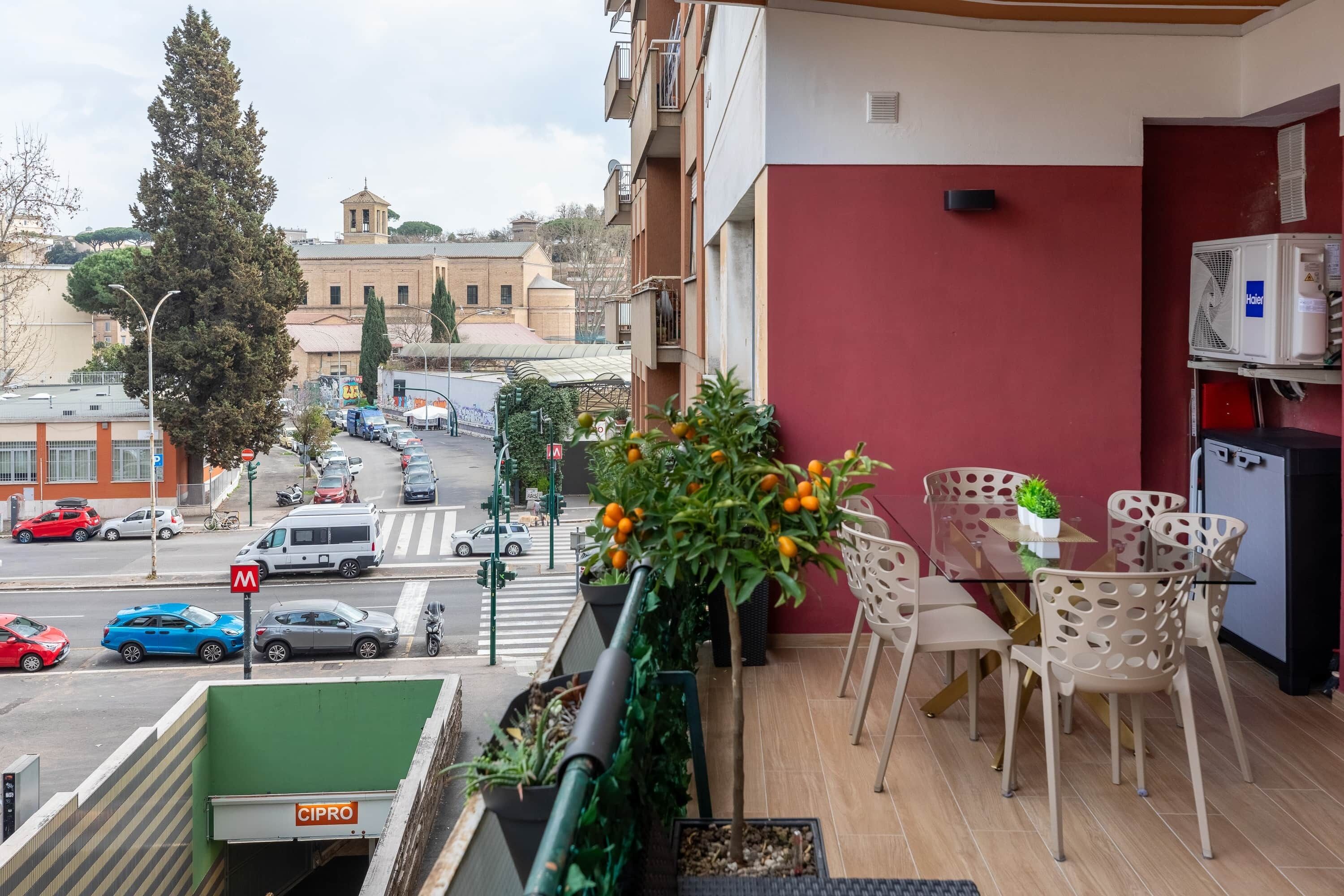 Un balcon avec une table en verre, des chaises, un pot de fleurs et un mur rouge.