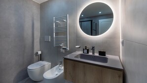 Et moderne badeværelse med toilet, bidet, vask og et rundt spejl med lys.