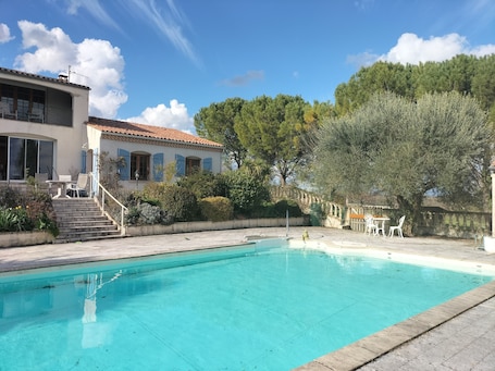 A swimming pool with a clear blue water, surrounded by a stone deck. There is a building with a tiled roof and blue shutters in the background.