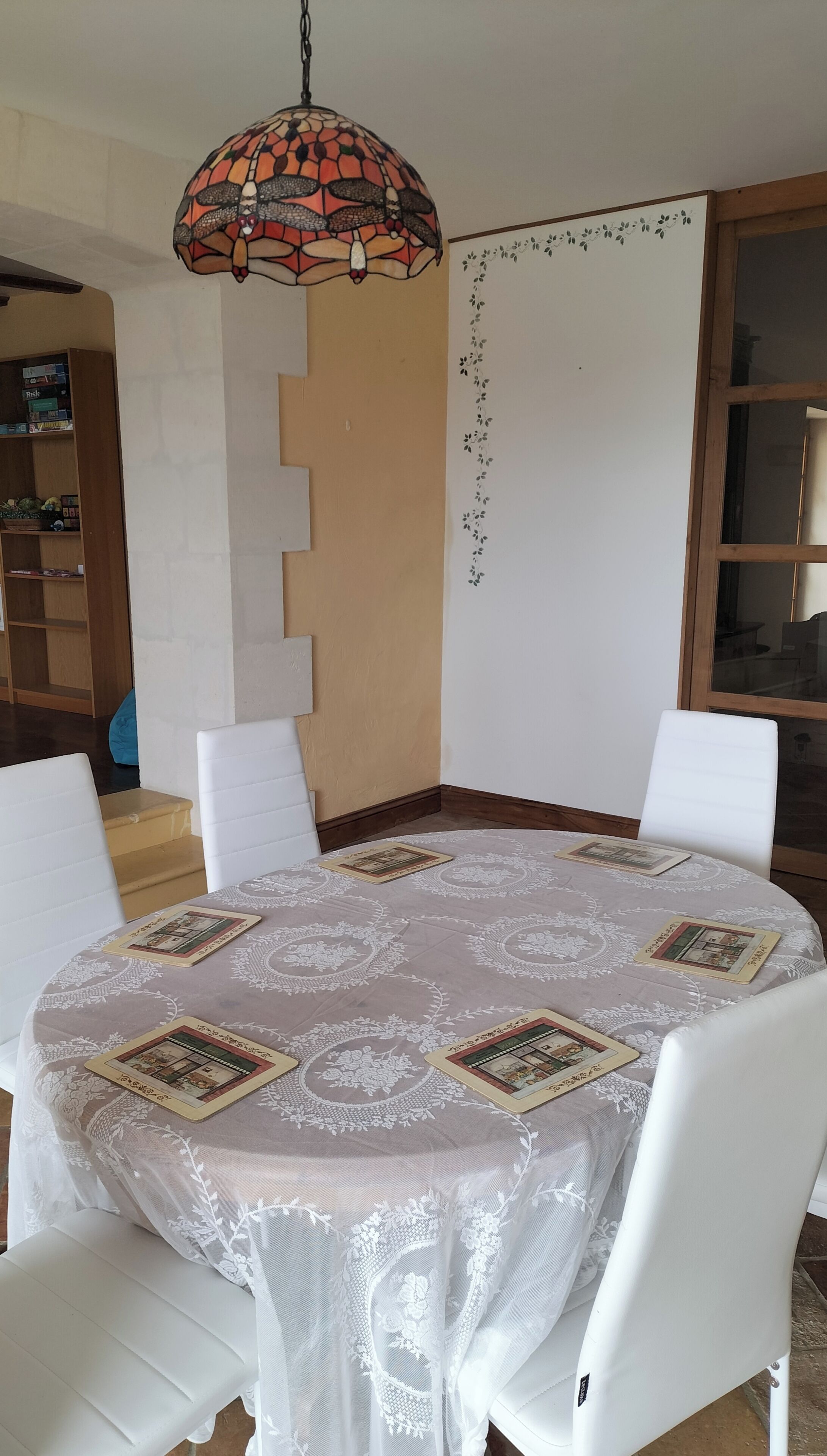 Een eetgedeelte met een ronde tafel, witte stoelen, een hanglamp van mozaïekglas en een wand met bloemenmotief.