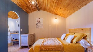 Ein Schlafzimmer mit Holzbalkendecke, einem Bett mit gelber Bettwäsche, einem Schminktisch, einem Bild an der Wand und einem kleinen Tisch mit Lampe.
