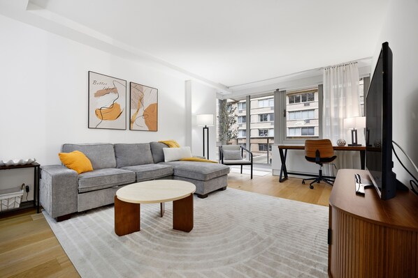 Ein modernes Wohnzimmer mit einem grauen Ecksofa, einem runden Couchtisch, einem Holztisch und einem großen Flachbildfernseher.
