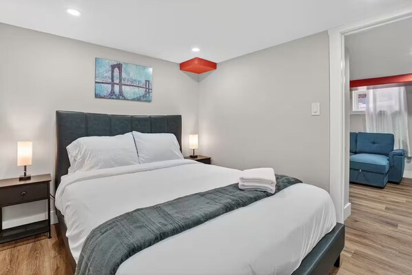 A modern hotel room with a large bed, a nightstand, a bedside lamp, a painting of a bridge, and a blue sofa in the corner. - Walkable to DT | Suits Large Grps | 2 Parking spot (Kitchener)