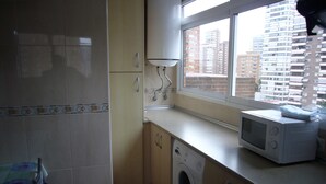 Une cuisine compacte équipée d’un lave-linge, d’un four à micro-ondes et d’une fenêtre donnant sur des immeubles de grande hauteur.