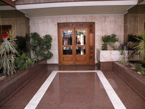 Entrada de hotel com piso de mármore, porta dupla de madeira e plantas em vasos de ambos os lados.